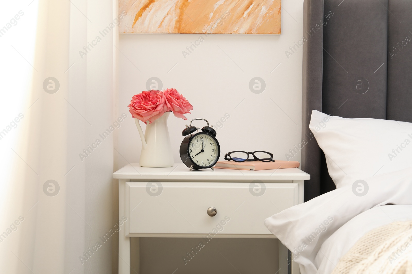 Bedside table with alarm clock, flowers and glasses near bed indoors Photo of Bedside table with alarm clock, flowers and glasses near bed indoors