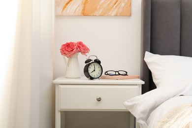 Photo of Bedside table with alarm clock, flowers and glasses near bed indoors