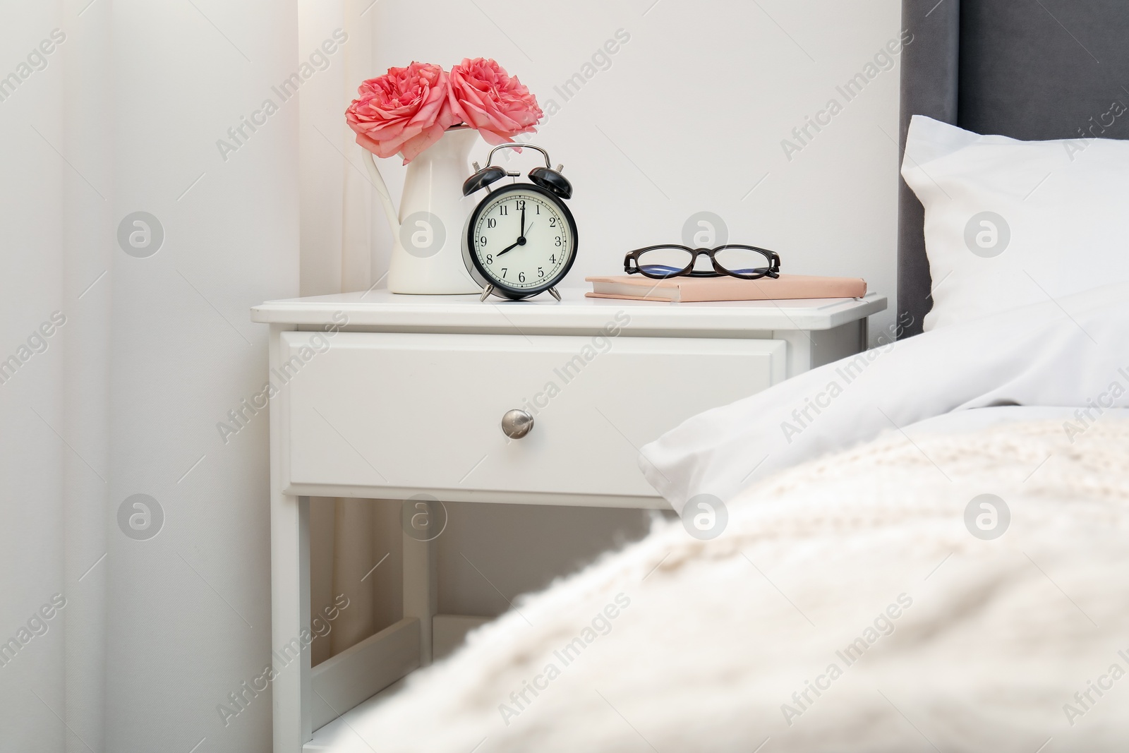 Bedside table with alarm clock, flowers and glasses near bed indoors Photo of Bedside table with alarm clock, flowers and glasses near bed indoors