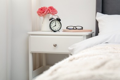 Photo of Bedside table with alarm clock, flowers and glasses near bed indoors
