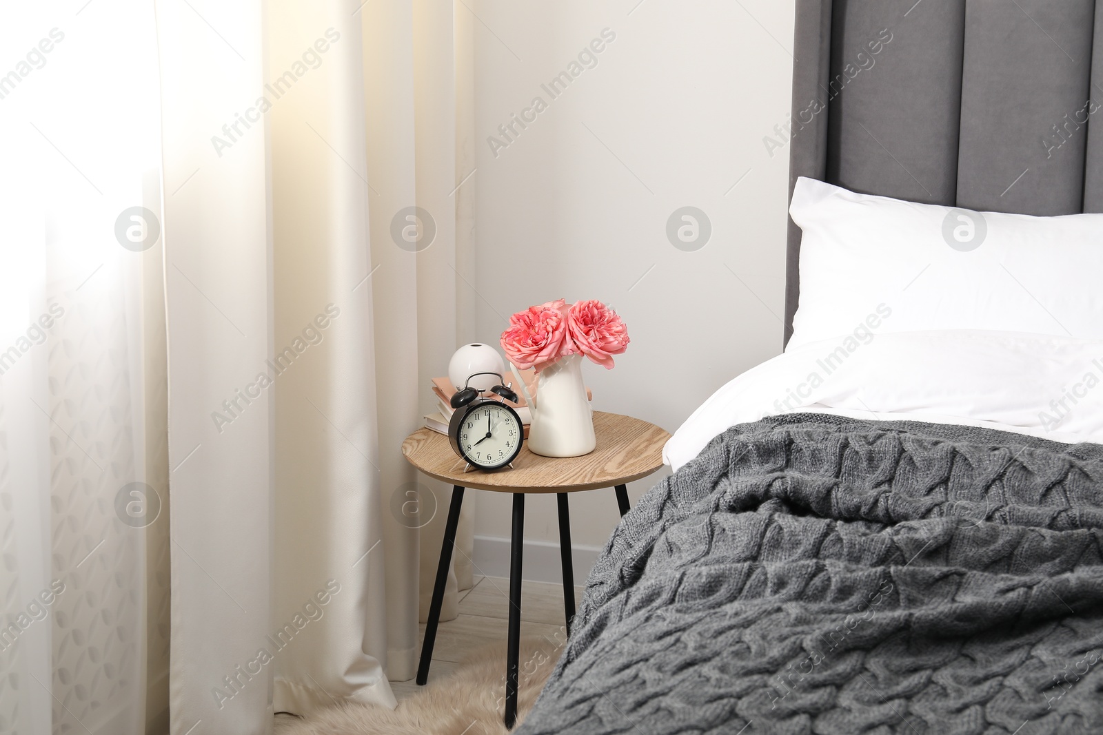 Bedside table with alarm clock, flowers and books near bed indoors Photo of Bedside table with alarm clock, flowers and books near bed indoors
