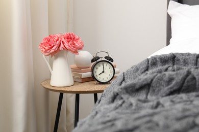Photo of Bedside table with alarm clock, flowers and books near bed indoors