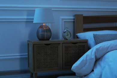 Photo of Bedside table with lamp and alarm clock near bed indoors