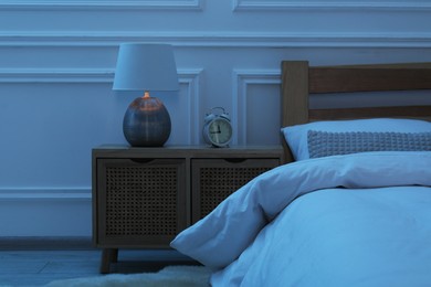 Photo of Bedside table with lamp and alarm clock near bed indoors