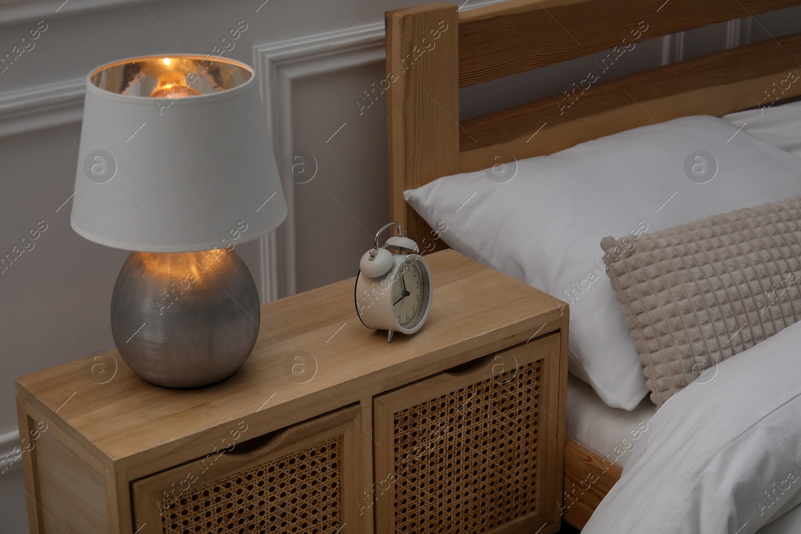 Bedside table with lamp and alarm clock near bed indoors Photo of Bedside table with lamp and alarm clock near bed indoors