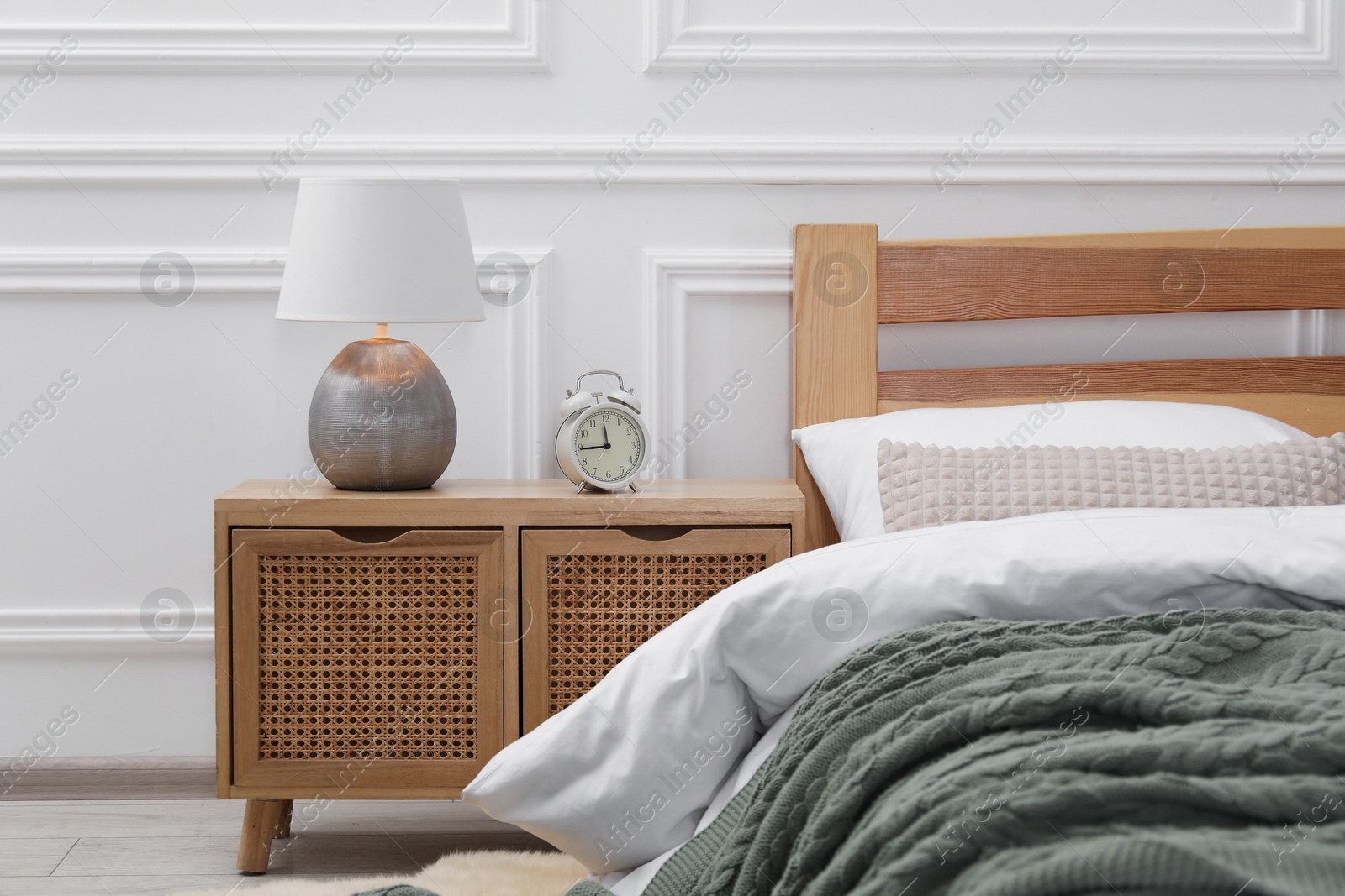 Bedside table with lamp and alarm clock near bed indoors Photo of Bedside table with lamp and alarm clock near bed indoors