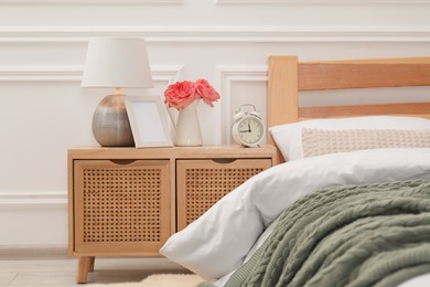 Photo of Bedside table with lamp, alarm clock and flowers near bed indoors