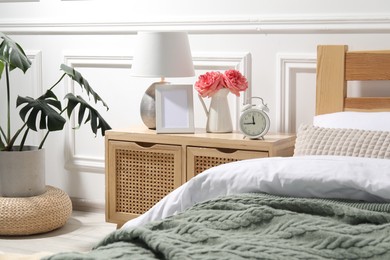 Photo of Bedside table with lamp, alarm clock and flowers near bed indoors