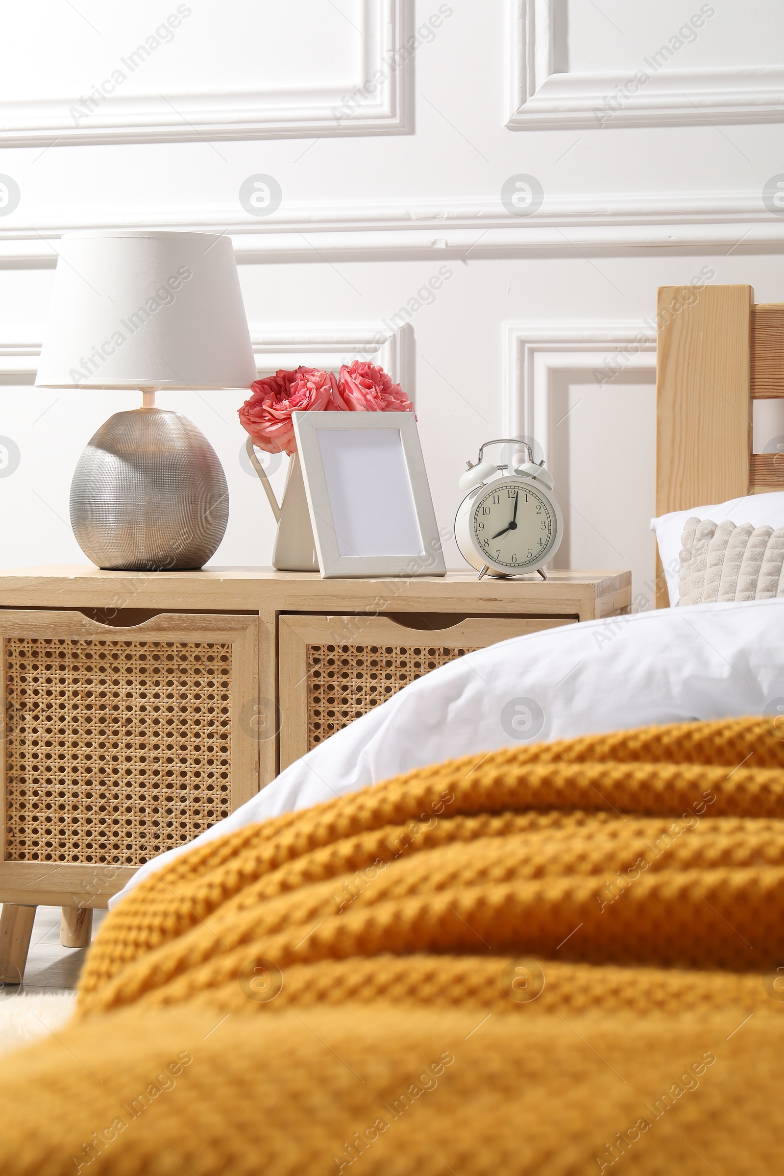 Bedside table with lamp, alarm clock and flowers near bed indoors Photo of Bedside table with lamp, alarm clock and flowers near bed indoors