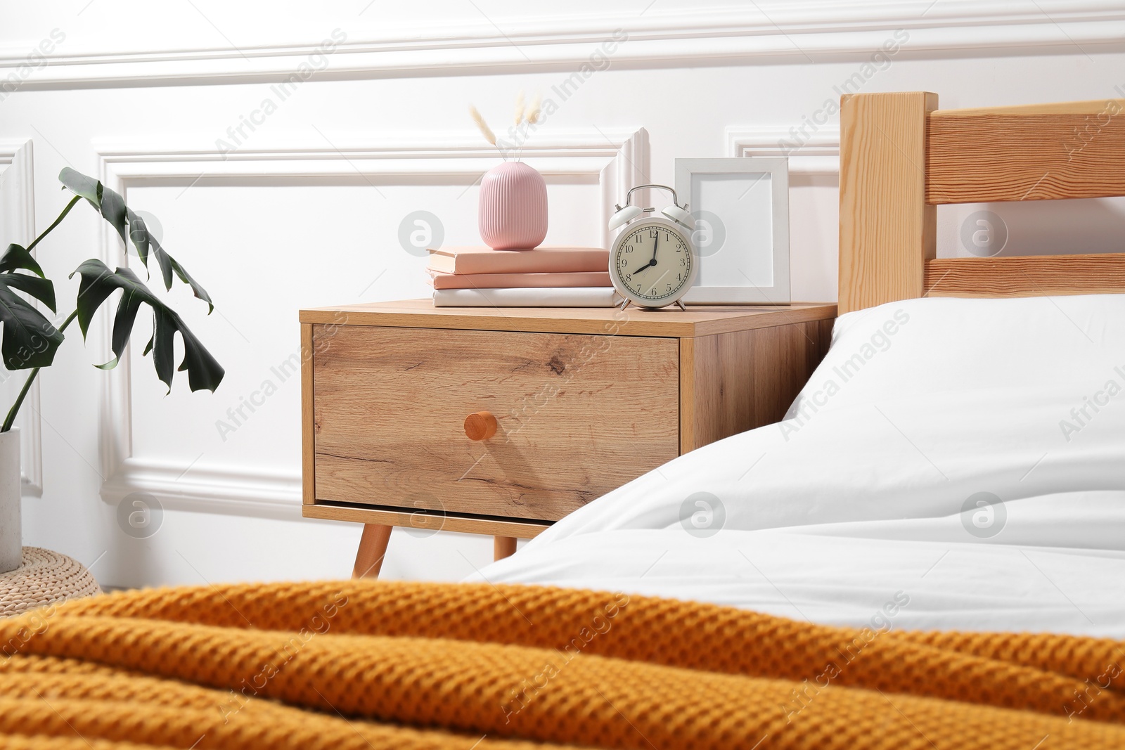 Bedside table with alarm clock, books and decor near bed indoors Photo of Bedside table with alarm clock, books and decor near bed indoors
