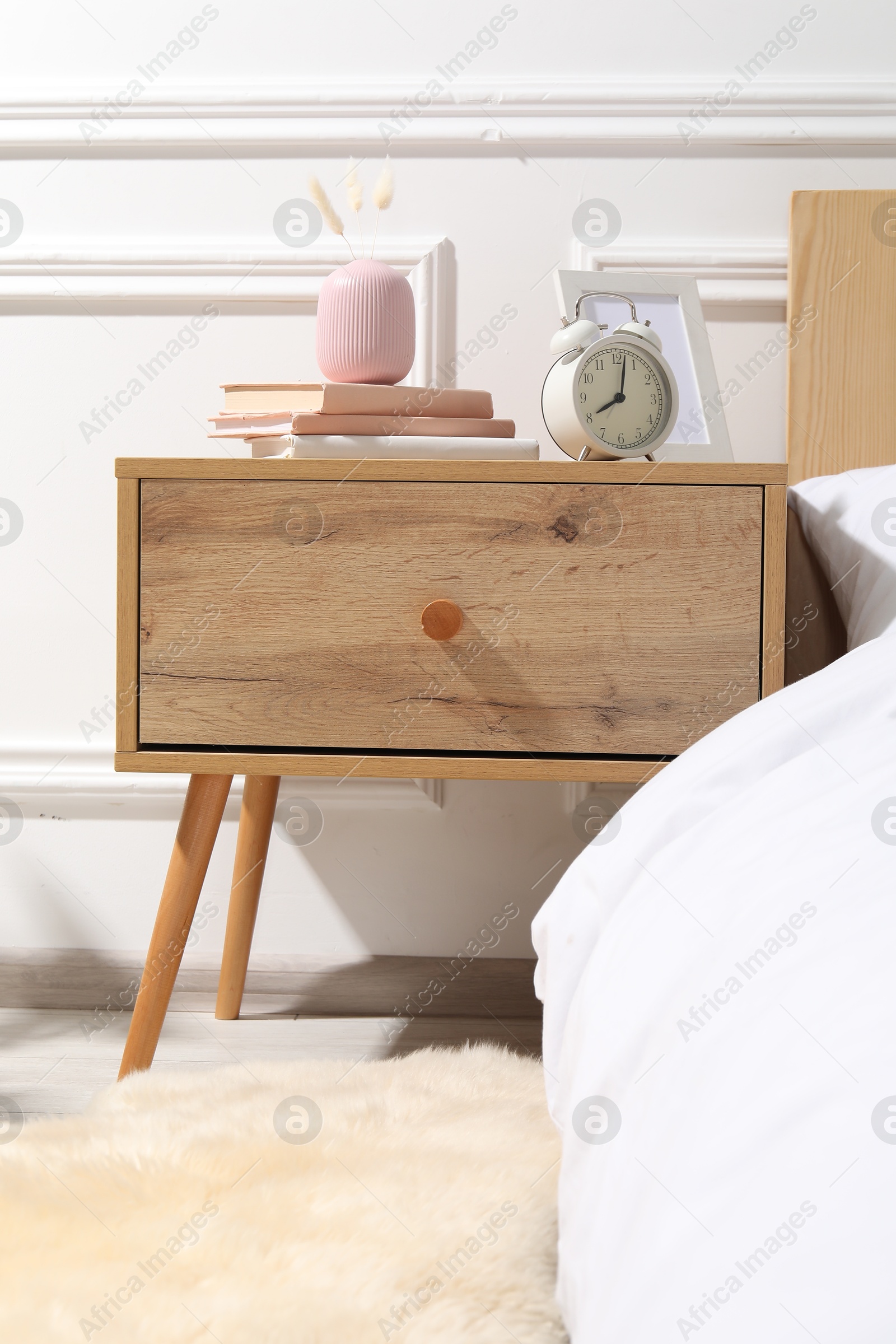 Bedside table with alarm clock, books and decor near bed indoors Photo of Bedside table with alarm clock, books and decor near bed indoors