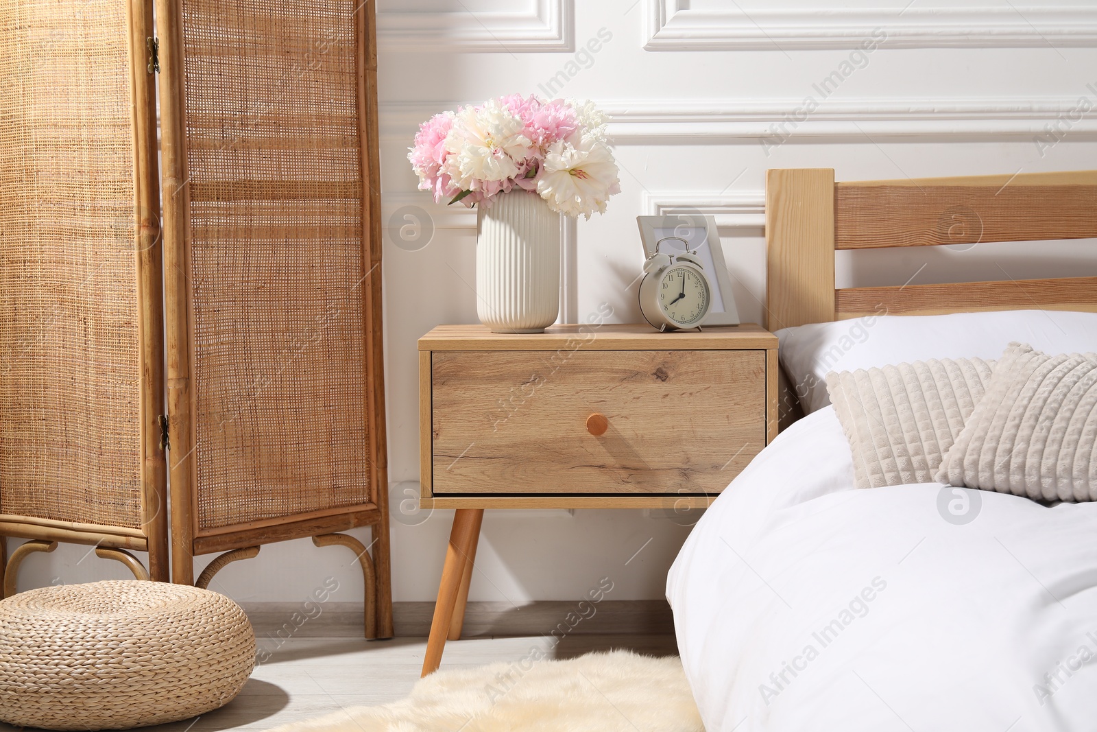 Bedside table with alarm clock and flowers near bed indoors Photo of Bedside table with alarm clock and flowers near bed indoors