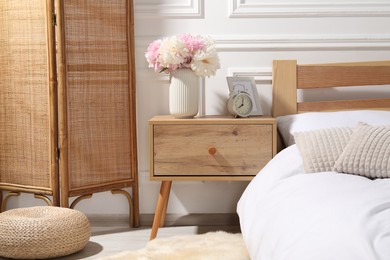 Photo of Bedside table with alarm clock and flowers near bed indoors