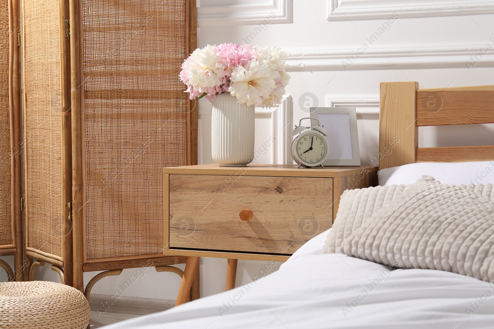 Bedside table with alarm clock and flowers near bed indoors Photo of Bedside table with alarm clock and flowers near bed indoors