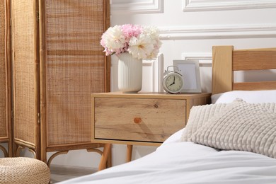 Photo of Bedside table with alarm clock and flowers near bed indoors