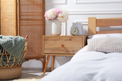Photo of Bedside table with alarm clock and flowers near bed indoors