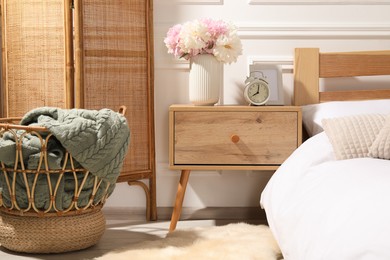 Photo of Bedside table with alarm clock and flowers near bed indoors