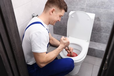Photo of Plumber unclogging toilet with hand plunger indoors