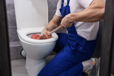 Photo of Plumber unclogging toilet with hand plunger indoors, closeup