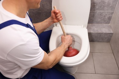 Photo of Plumber unclogging toilet with hand plunger indoors, closeup