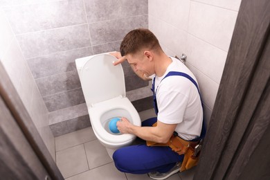 Photo of Plumber unclogging toilet with hand plunger indoors