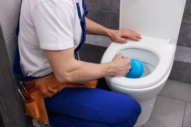 Photo of Plumber unclogging toilet with hand plunger indoors, closeup