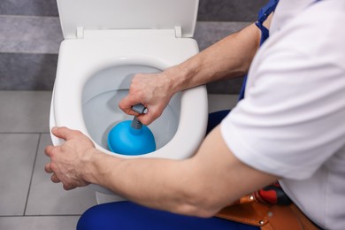 Photo of Plumber unclogging toilet with hand plunger indoors, closeup