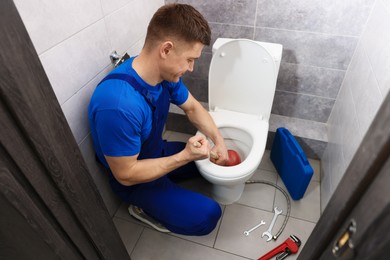 Photo of Plumber unclogging toilet with hand plunger indoors