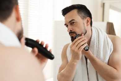 Photo of Handsome man shaving with trimmer near mirror at home