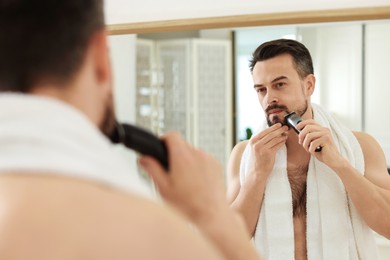 Photo of Handsome man shaving with trimmer near mirror at home