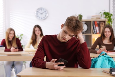 Photo of Cyber bullying. Upset teenage boy with smartphone at desk in classroom