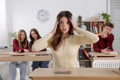 Photo of Cyber bullying. Upset teenage girl with smartphone and schoolmates pointing at her in classroom, selective focus