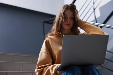 Photo of Cyber bullying. Upset teenage girl with laptop on stairs indoors. Space for text