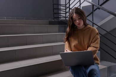 Photo of Cyber bullying. Upset teenage girl with laptop on stairs indoors. Space for text
