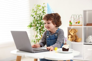 Photo of Birthday celebration. Little boy with cupcake having video call via laptop at table indoors