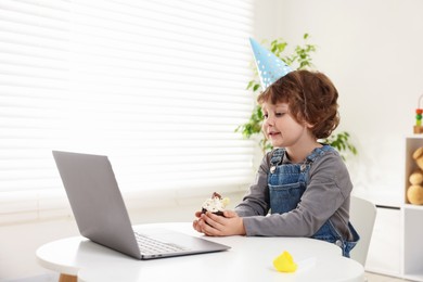 Photo of Birthday celebration. Little boy with cupcake having video call via laptop at table indoors. Space for text