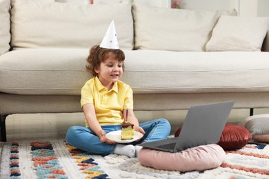 Photo of Birthday celebration. Little boy with piece of cake having video call via laptop at home