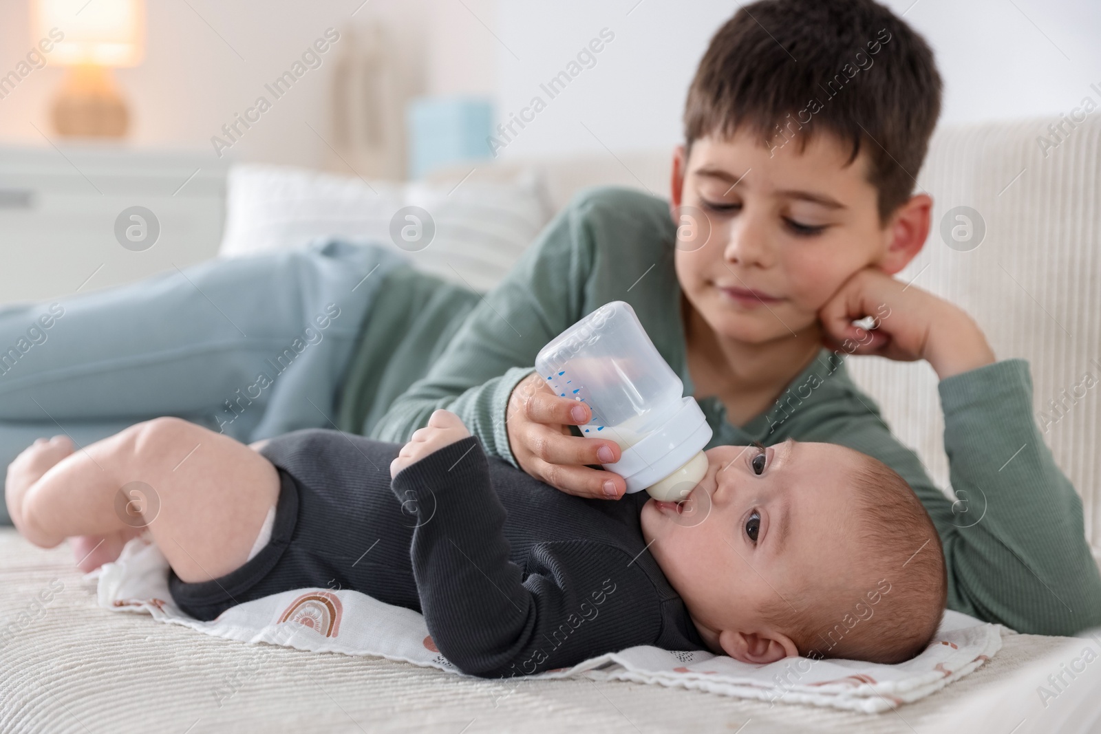 Little boy feeding his newborn brother with bottle on sofa at home Photo of Little boy feeding his newborn brother with bottle on sofa at home