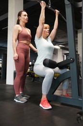 Photo of Senior woman training on lat pulldown machine supervised by professional trainer in gym