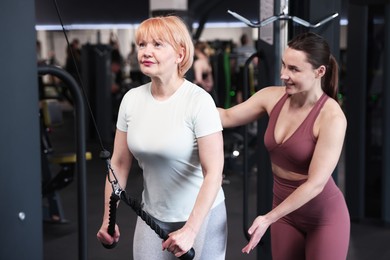 Photo of Senior woman training on pulley machine supervised by professional trainer in gym