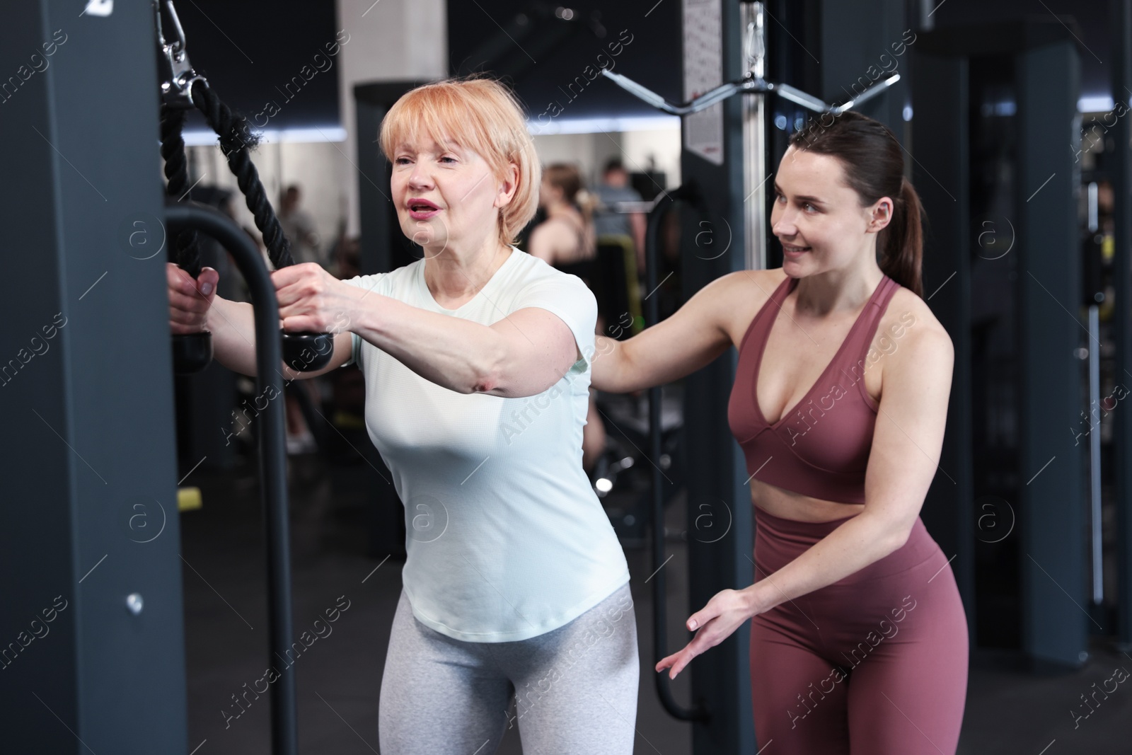 Senior woman training on pulley machine supervised by professional trainer in gym Photo of Senior woman training on pulley machine supervised by professional trainer in gym
