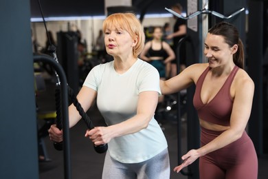Photo of Senior woman training on pulley machine supervised by professional trainer in gym