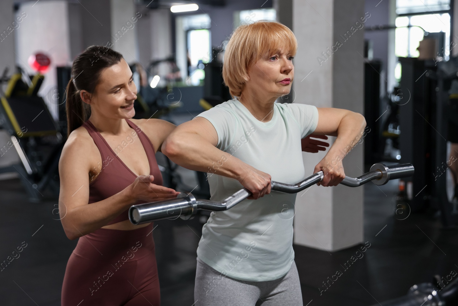 Senior woman exercising with EZ curl bar supervised by professional trainer in gym Photo of Senior woman exercising with EZ curl bar supervised by professional trainer in gym