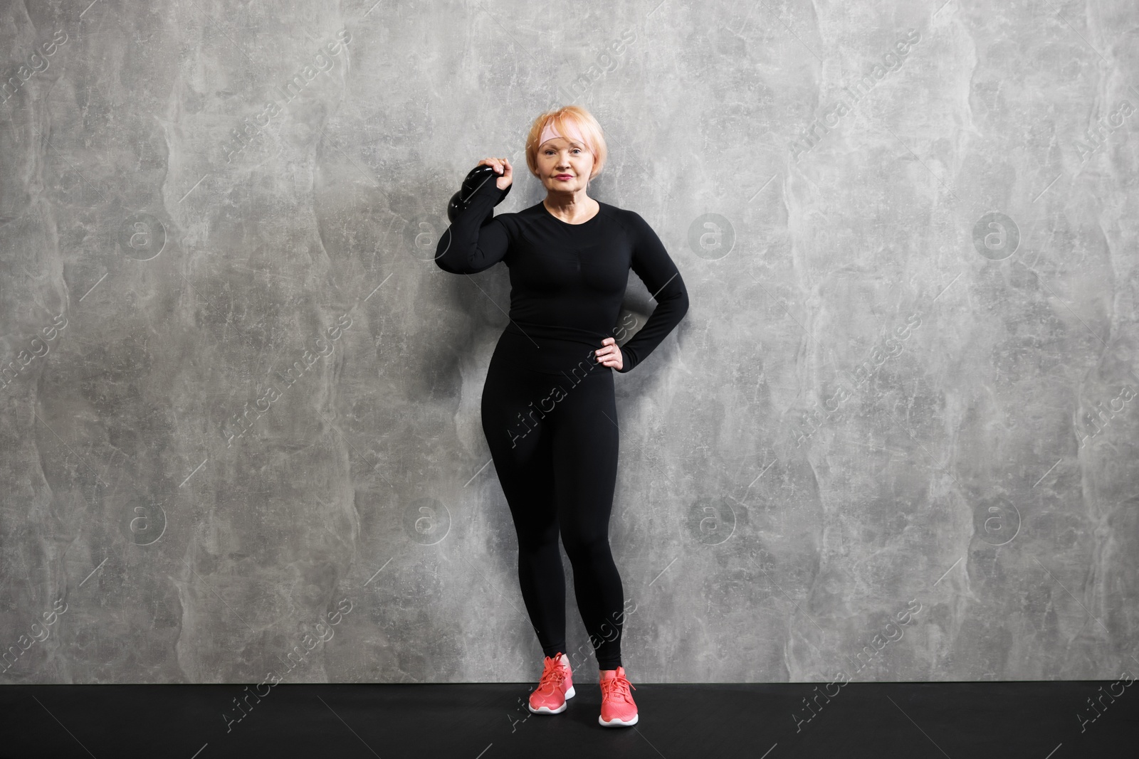 Senior woman with kettlebell near gray marble wall indoors Photo of Senior woman with kettlebell near gray marble wall indoors