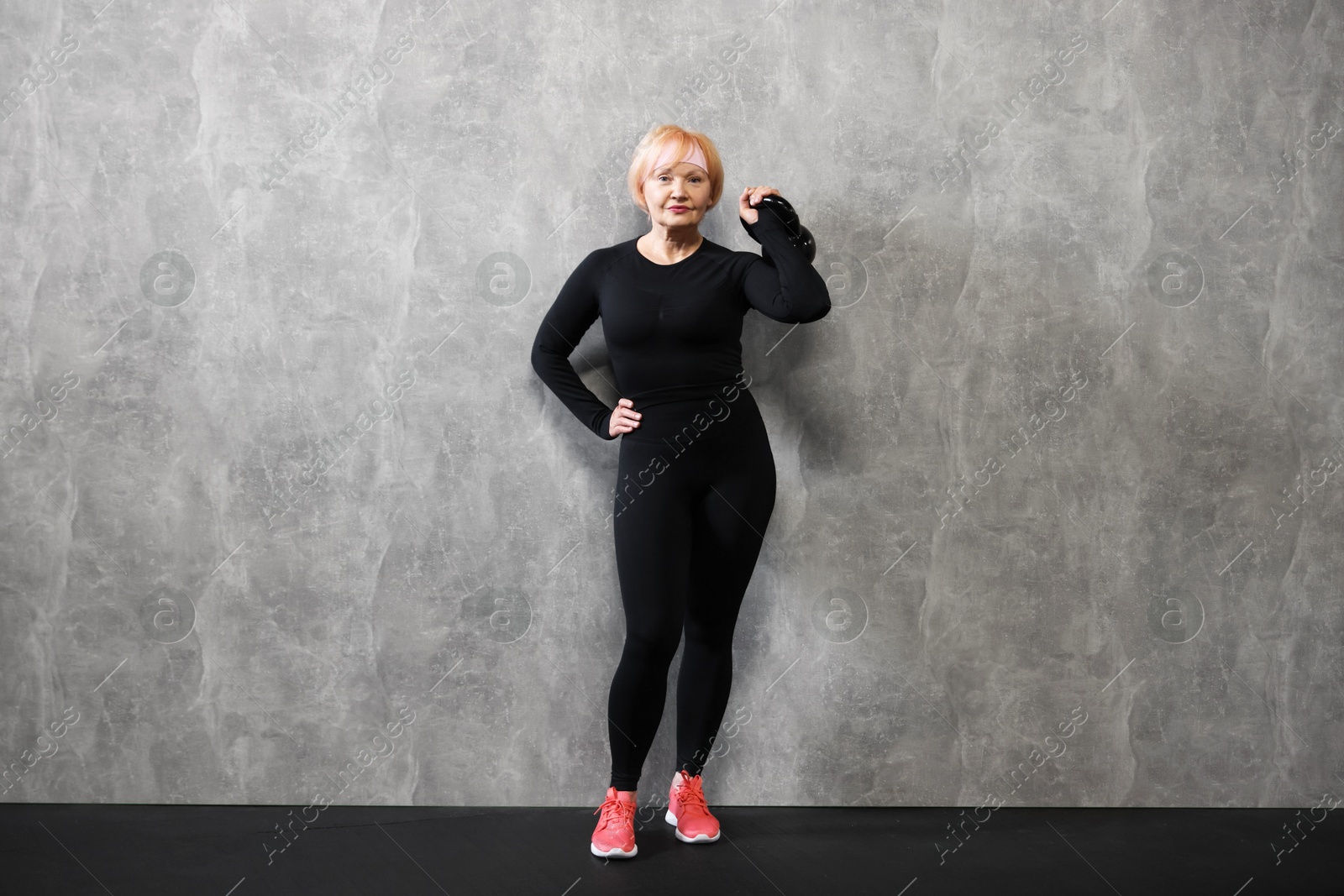Senior woman with kettlebell near gray marble wall indoors Photo of Senior woman with kettlebell near gray marble wall indoors