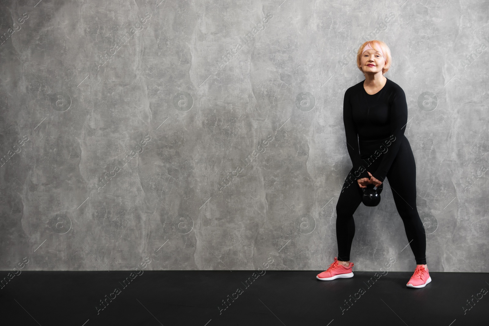 Senior woman with kettlebell near gray marble wall indoors, space for text Photo of Senior woman with kettlebell near gray marble wall indoors, space for text