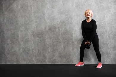 Photo of Senior woman with kettlebell near gray marble wall indoors, space for text