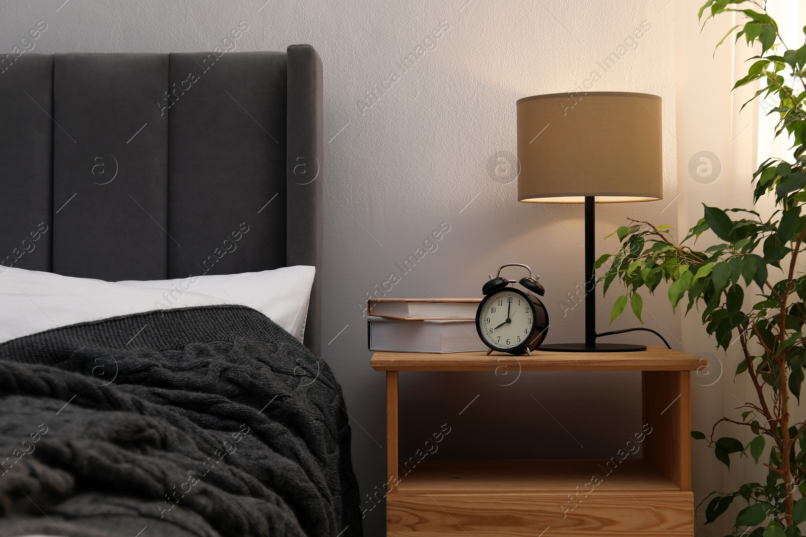Bedside table with lamp, alarm clock and books near bed indoors. Interior design Photo of Bedside table with lamp, alarm clock and books near bed indoors. Interior design