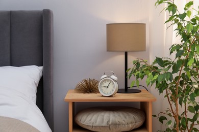 Photo of Bedside table with lamp, alarm clock and decor near bed indoors. Interior design