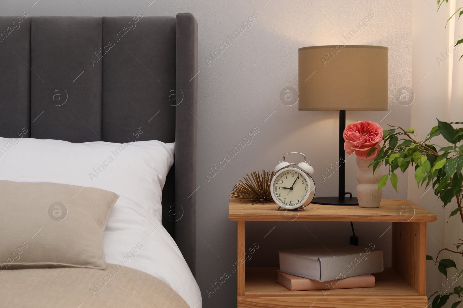 Bedside table with lamp, alarm clock, books and decor near bed indoors. Interior design Photo of Bedside table with lamp, alarm clock, books and decor near bed indoors. Interior design